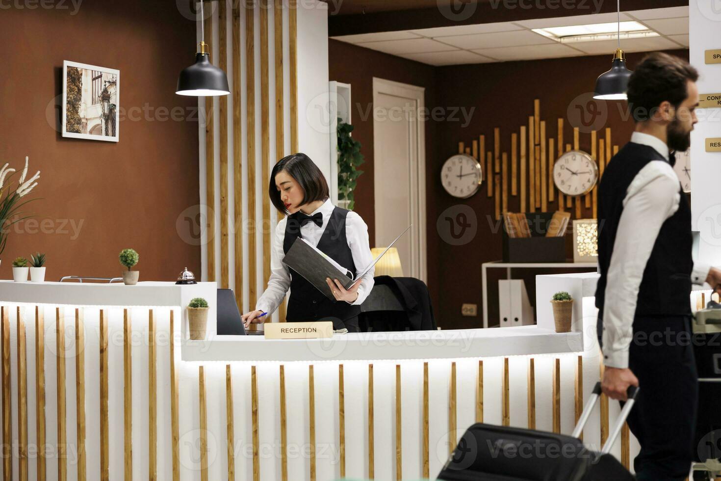 Receptionist reviewing check in reports, looking at record files before helping guests with accomodation in luxury hotel. Asian woman working at front desk in hotel lobby reception. photo