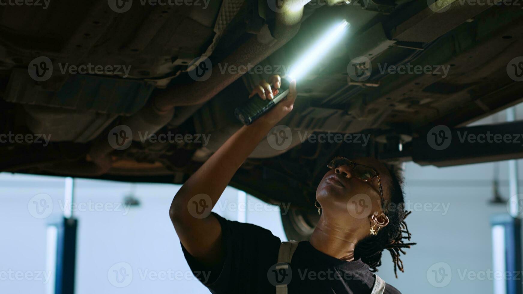 Licensed mechanic standing underneath suspended car in garage workspace, using work light to check for damages and tablet to order new replacing parts after finding issues during annual maintenance photo