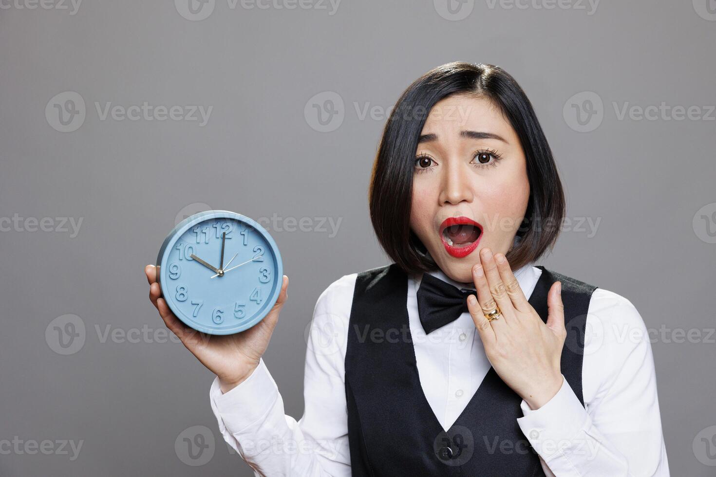 Shocked overslept asian waitress holding alarm clock and looking at camera while covering open mouth. Young anxious woman receptionist showing time while running late portrait photo