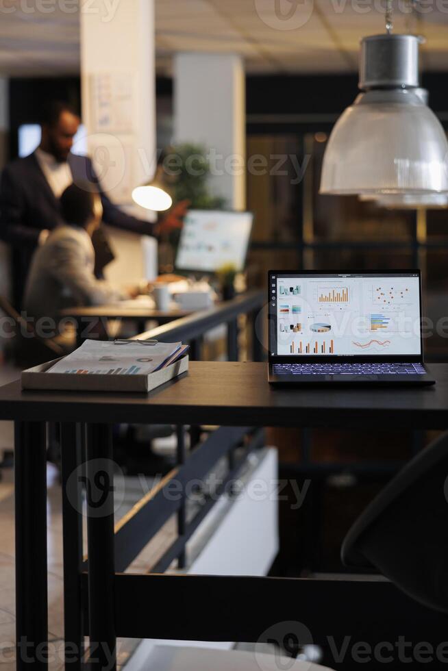 Laptop computer with financial graphs on screen, standing on table in startup office, coworkers discussing company strategy in background. Employees working late at night in startup office photo