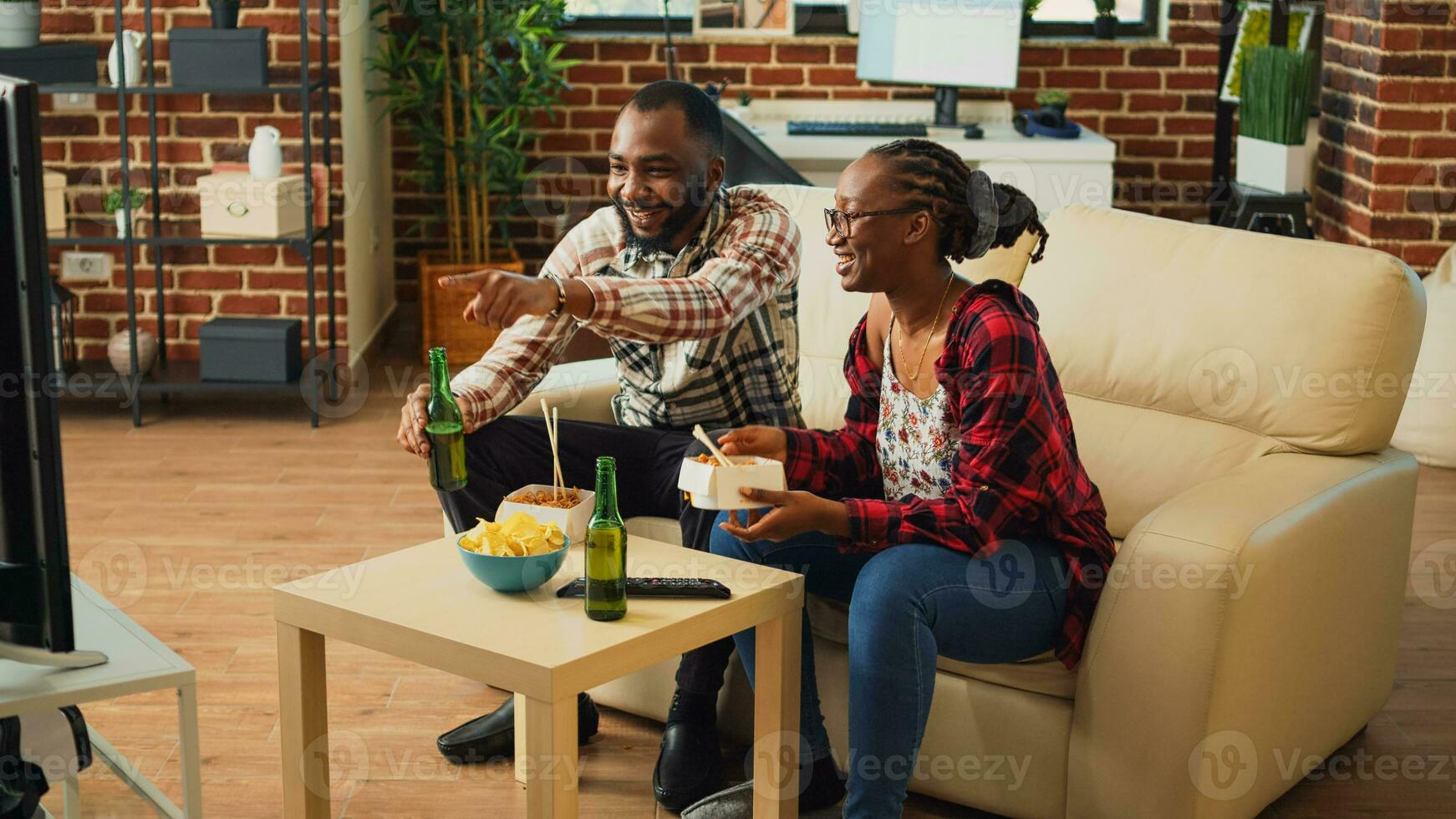 Happy life partners eating asian meal from delivery, serving noodles with chopsticks and watching comedy movie on tv. Modern couple in relationship enjoying show and food with alcohol. photo