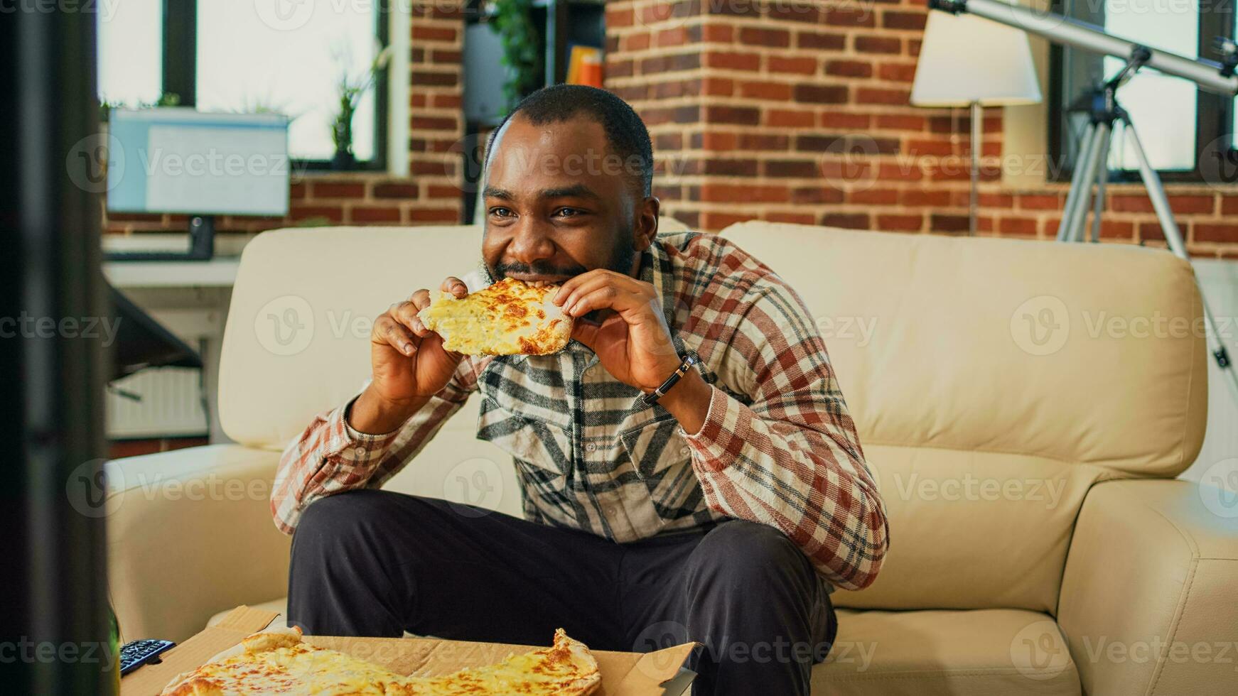 Happy guy eating slice of pizza and drinking beer at home, enjoying takeout food and watching television. Young man serving takeaway dinner and alcohol, watch movie in living room. photo