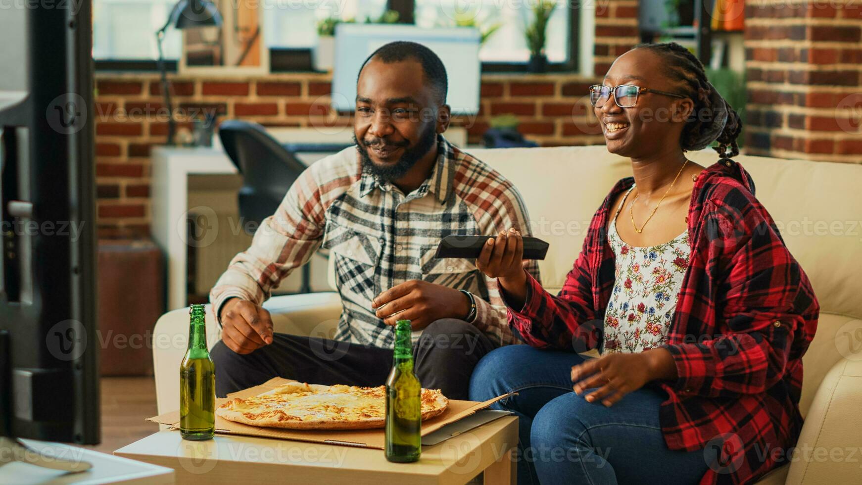 Cheerful man and woman eating slices of pizza at home, being relaxed together watching movie on television. Young people in relationship enjoying delivery meal and beer bottles. photo