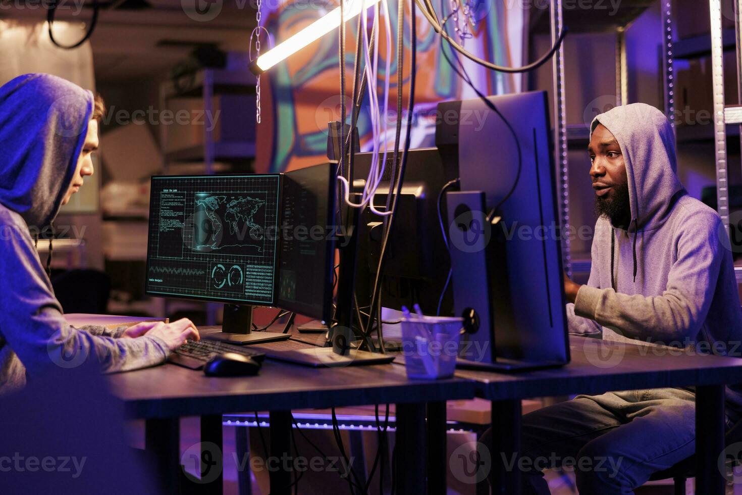 Two hackers planning phishing cyberattack while coding ransomware on computers. Asian and african american men in hoods developing malicious software in dark abandoned warehouse photo