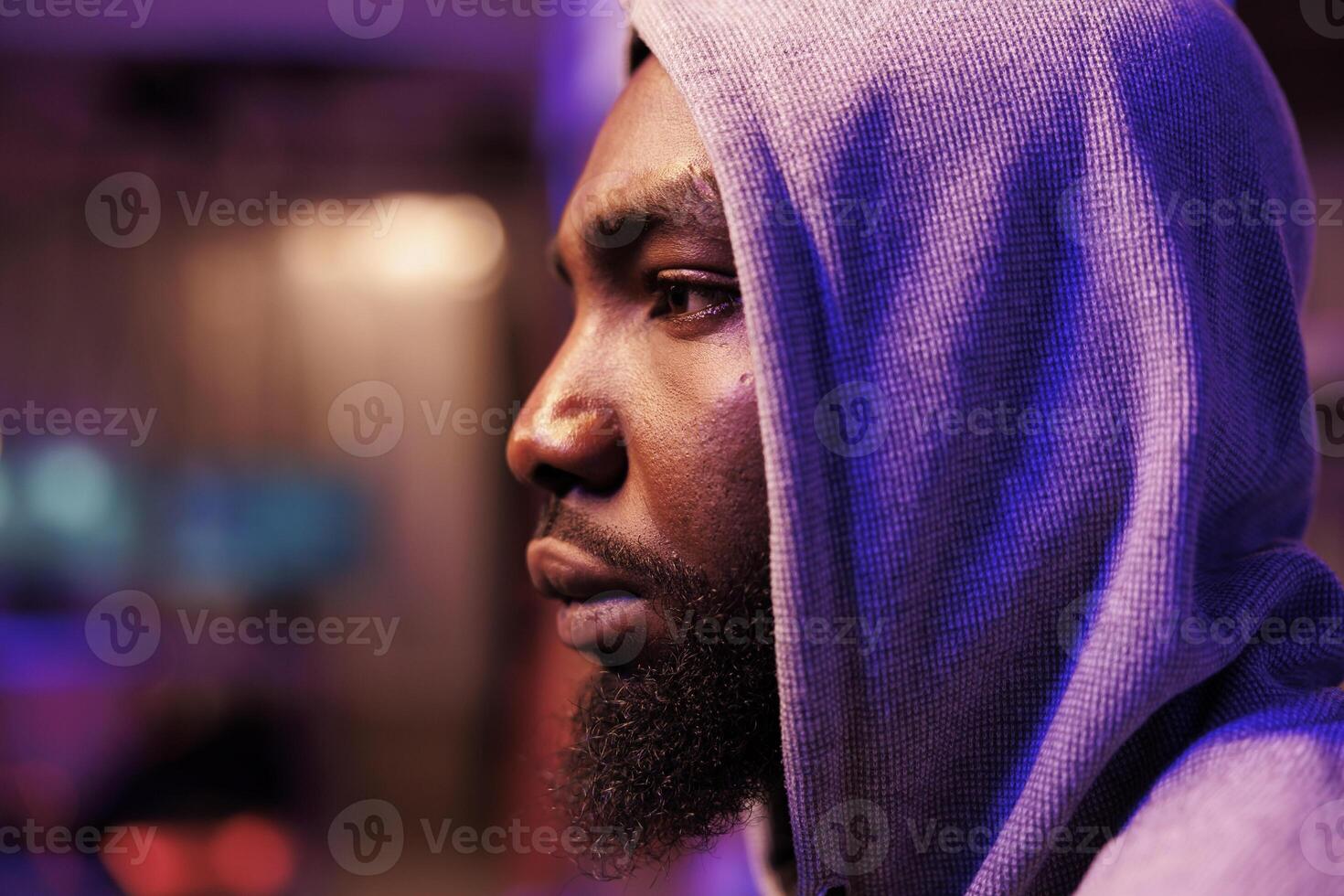 Concentrated african american man in hood breaking into computer system in dark room. Focused young hacker coding internet virus and ransomware in hideout with neon light close up photo