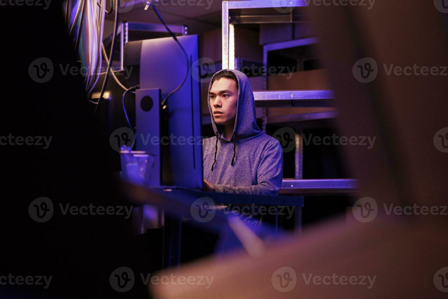 Asian hacker working on ransomware to break into government server and steal sensitive data. Young man in hood coding illegal malicious software and programming spyware at night photo