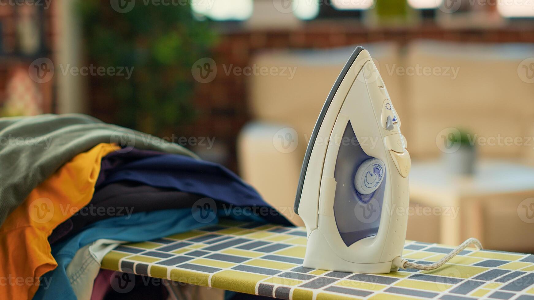 Iron tool placed on ironing board with pile of washed clothing, electric appliances used to do housekeeping work. Domestic chores with iron to smooth out creases on laundry. Close up. photo
