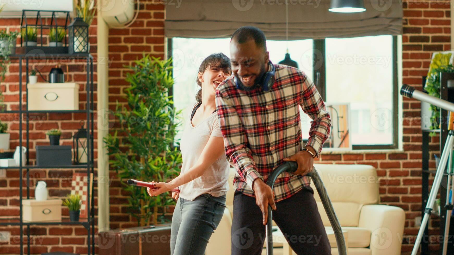 Young life partners vacuuming floors and showing dance moves at home, having fun doing housework. Cheerful modern man and woman dancing and singing on music, acting silly. Handheld shot. photo