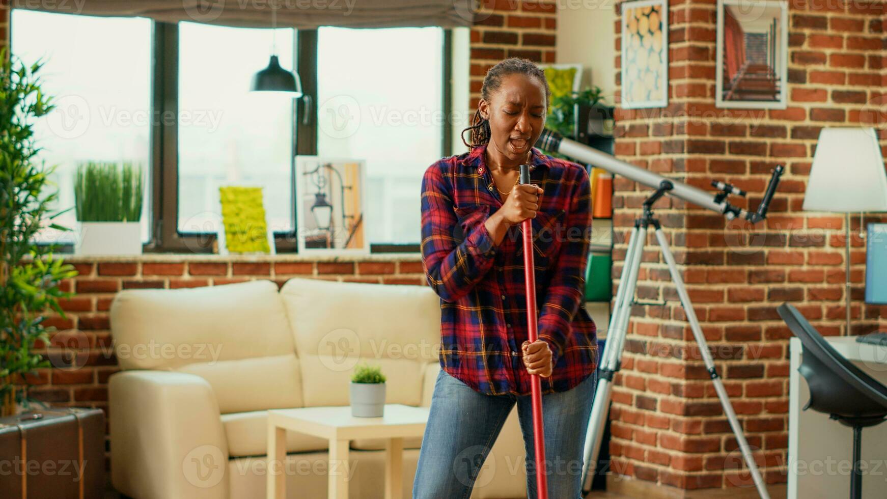 African american housewife listening to music and mopping apartment floors, using mop and washing solution. Young happy woman dancing and singing, having fun with spring cleaning. photo