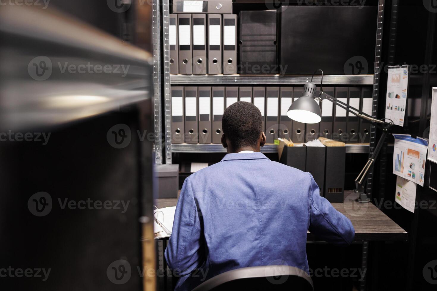 Bookkeeper sitting at desk in storage room, working at bureaucracy record, reading files with administrative report. African american manager analyzing accountancy documents in corporate depository photo
