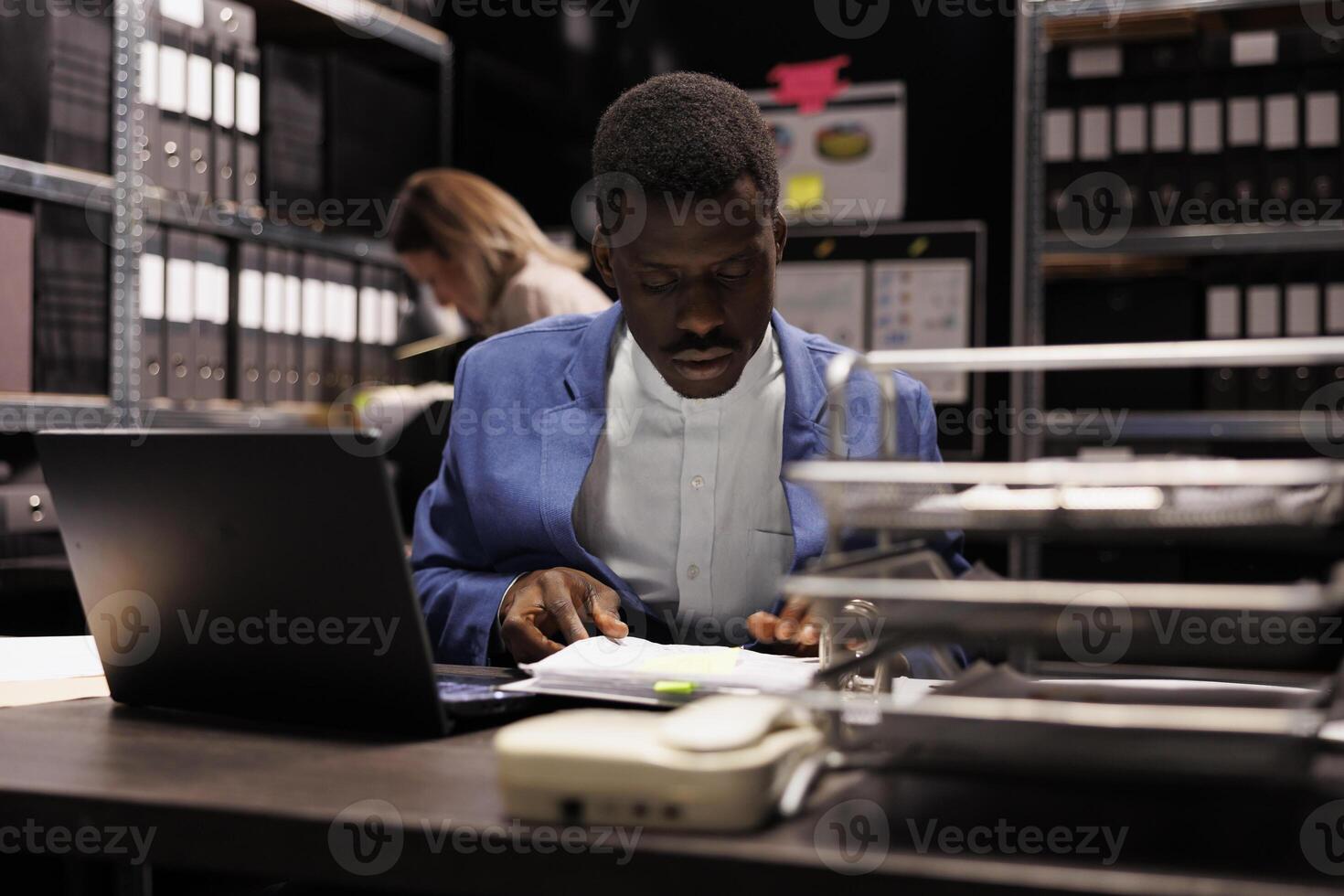 African american investigator analyzing police evidence, working overhours at criminal case in arhive room. Private detective checking crime scene evidence, searching for clues. Criminology concept photo