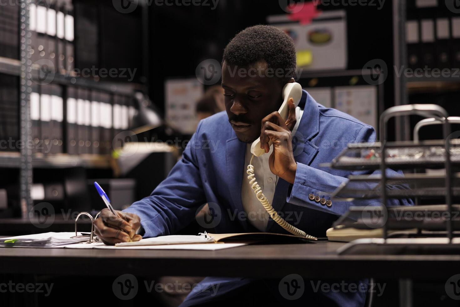African american police officer talking with remote detective using landline phone, discussing confidential clue. Private inspector analyzing crime scene evidence trying to solve case in arhive room photo