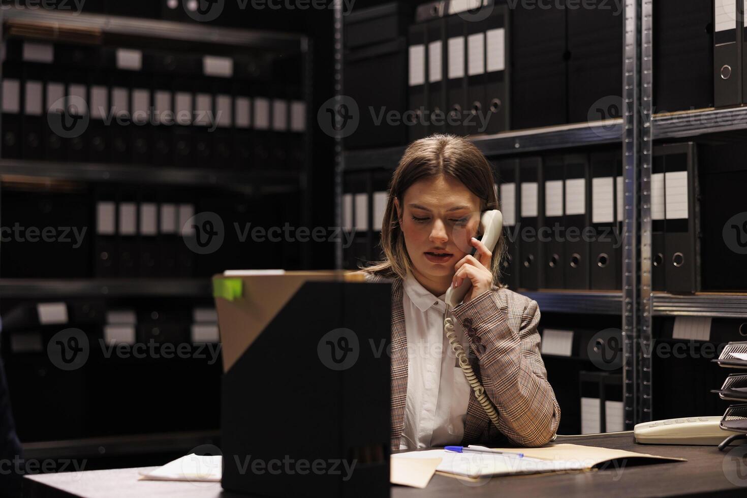 Bookkeeper discussing accountancy report with manager using landline phone, working overhours at management research in storage room. Repository worker checking bureaucracy record photo