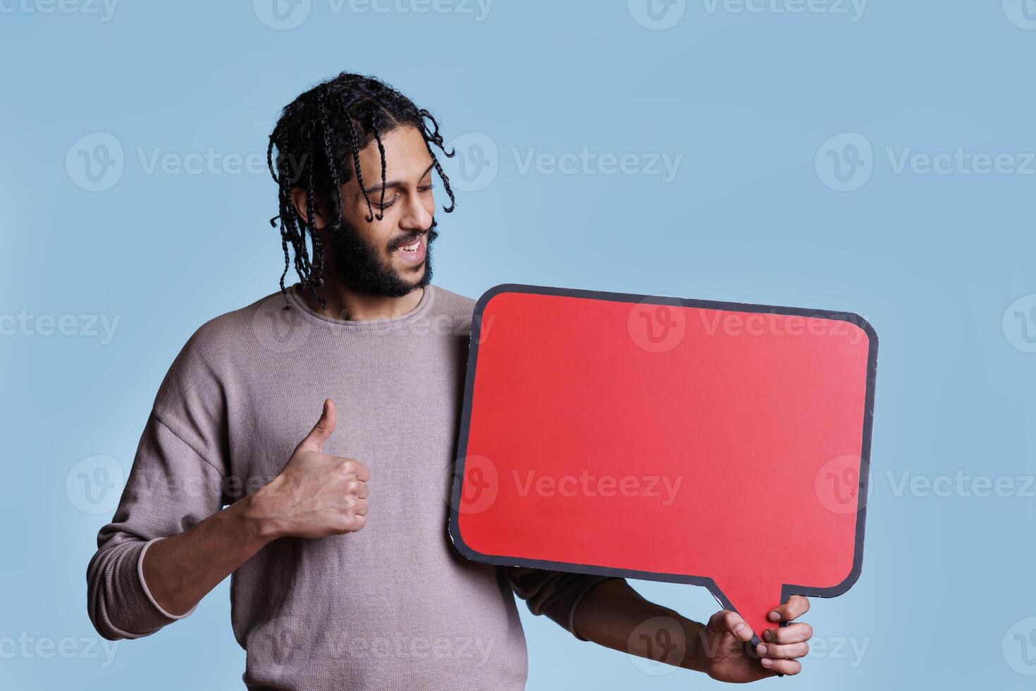 Smiling arab man holding empty red speech bubble and showing thumb up. Arabian person with happy facial expression standing with blank dialog frame for advertising message photo