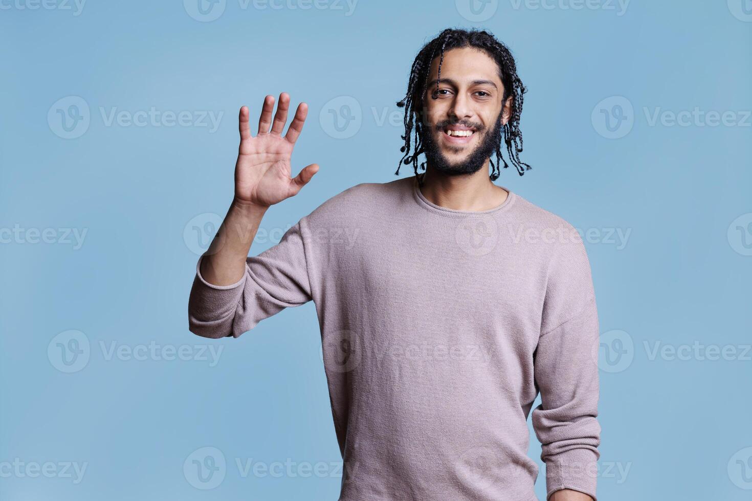 Friendly smiling arab man waving hello and looking at camera with positive emotions. Cheerful arabian handsome model wearing casual clothes raising hand for greeting portrait photo