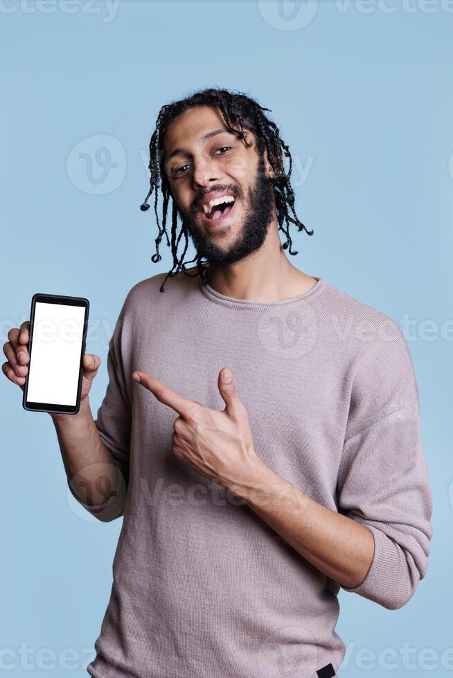 Smiling arab man presenting mobile phone with white screen for application advertisement and looking at camera. Cheerful advertiser pointing with finger at smartphone with empty display for text photo