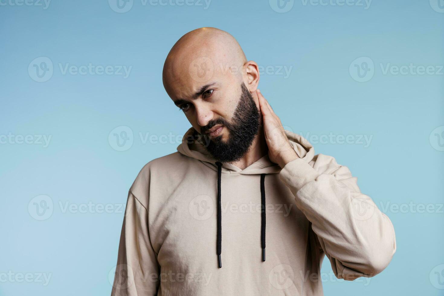 Arab man grimacing in discomfort while holding neck back and suffering from spasm. Tired young adult bald bearded person with exhausted facial expression rubbing sore muscles photo