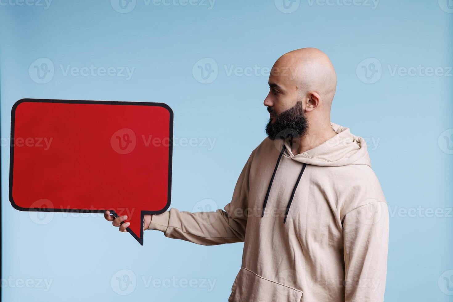 Arab man advertising product with empty red banner, showing frame for text. Young person holding blank dialog bubble shape with cope space for promotional message mock up photo