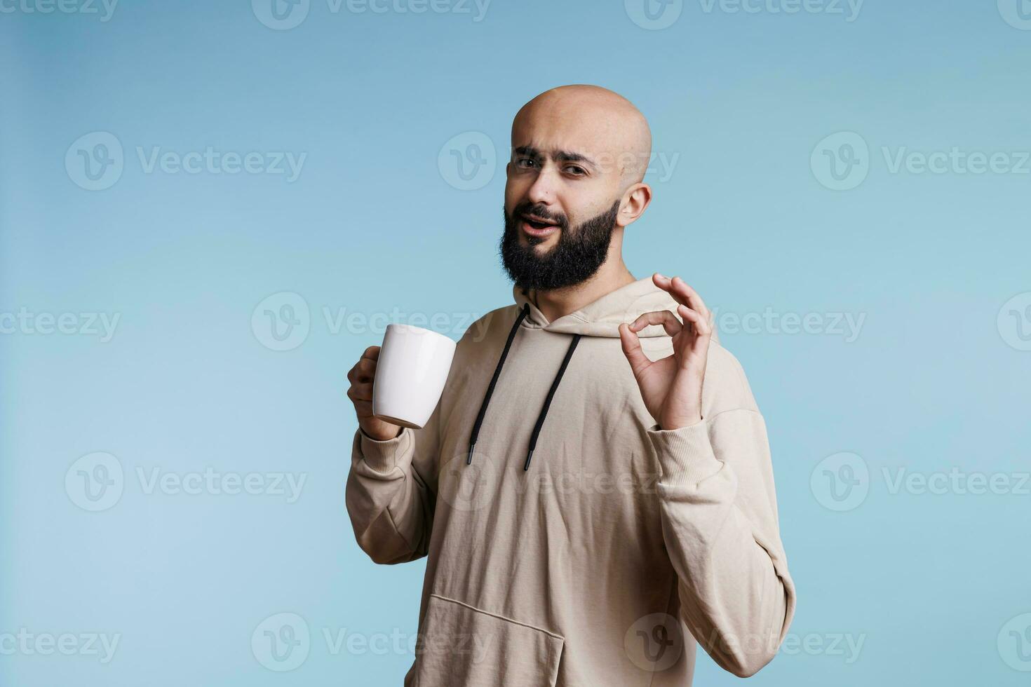 Arab man enjoying coffee and showing ok gesture with fingers portrait. Young person drinking tea from white mug and looking at camera with approval sign and satisfaction facial expression photo