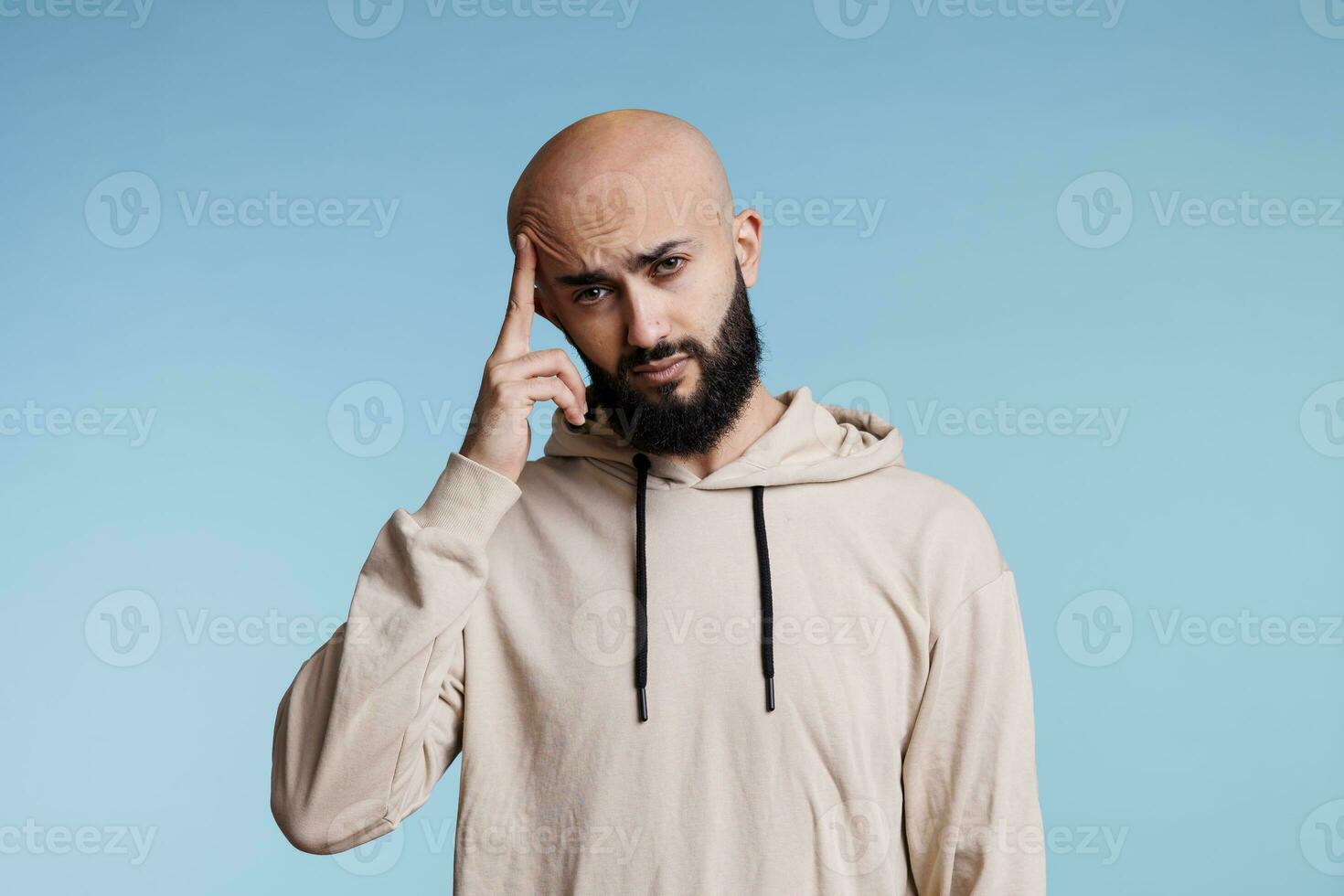 Arab man feeling pain of migraine while looking at camera with tired facial expression. Person rubbing temple while suffering from headache studio portrait on blue background photo