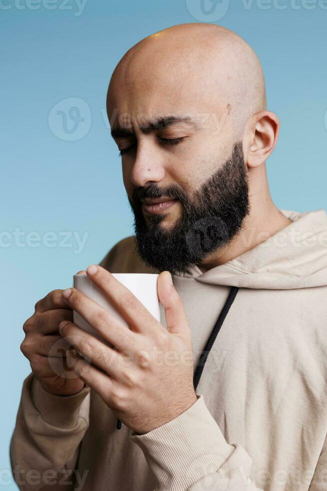 Relaxed arab man holding white cup in hands while enjoying coffee. Young thoughtful person wearing casual hoodie clothes drinking hot tea beverage from mug on blue background photo