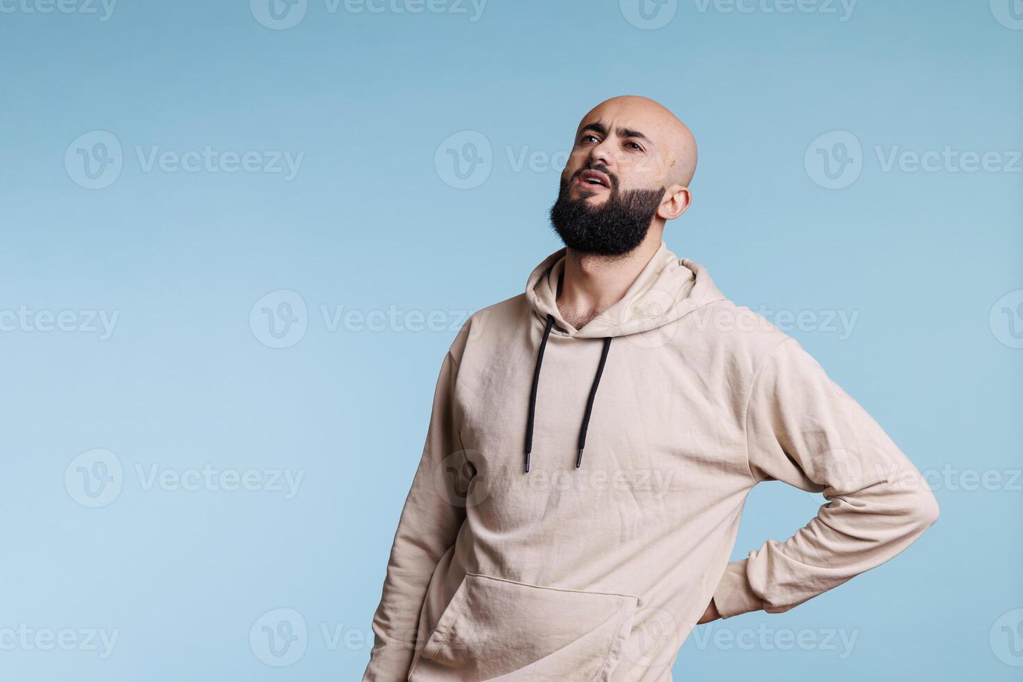 Young arab man holding hand on lower back while having spine problems. Bearded person looking upwards in pain and grimacing while suffering from backache and sore muscles cramps photo