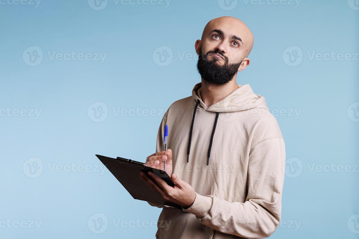 Thoughtful arab man holding clipboard and looking upwards with pensive facial expression. Puzzled person remembering while taking notes and writing list on blue background photo