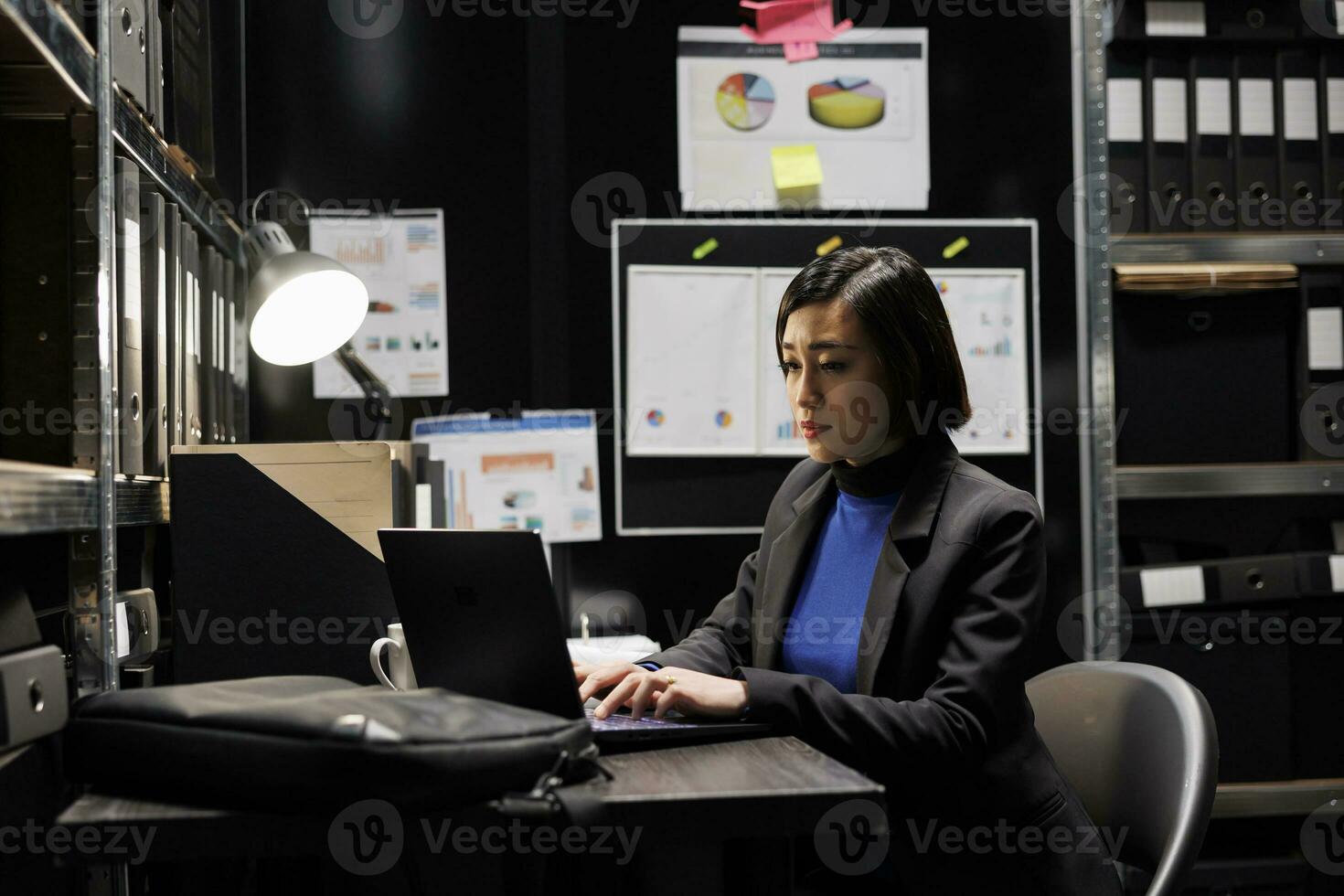 Professional businesswoman accountancy employee writing budget plan and accountancy strategy on laptop. Asian woman in file cabinet room filled with document folders and graphic charts photo