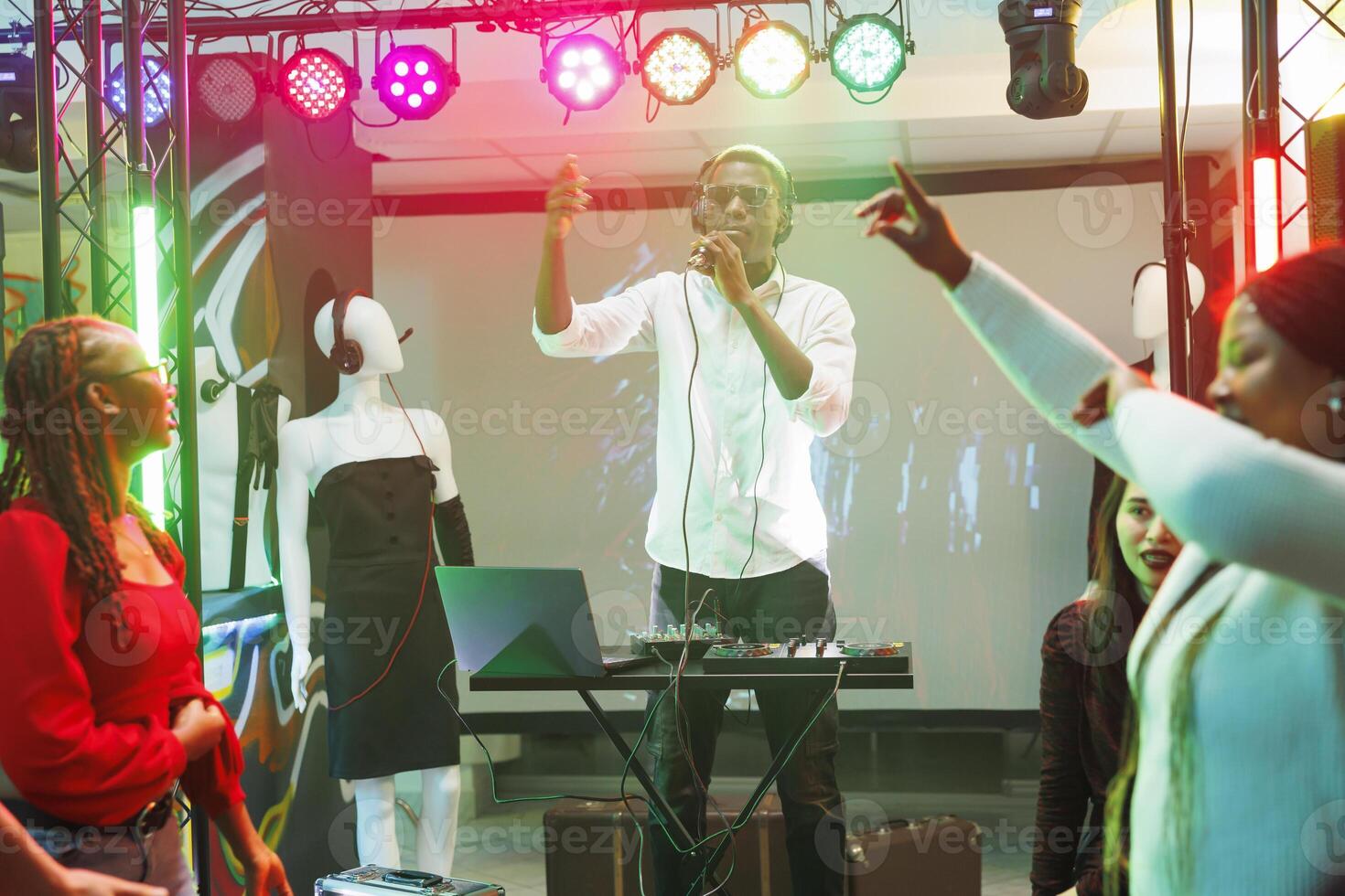 African american musician singing and playing using controller on stage in nightclub. Man dj with microphone performing at electronic music concert in club while crowd dancing photo