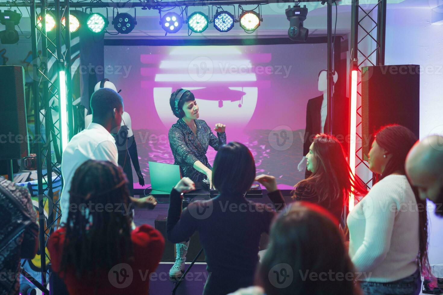 Energetic performer using dj station on stage to mix electronic music while crowd partying on dancefloor. Young people enjoying clubbing experience and dancing at concert photo