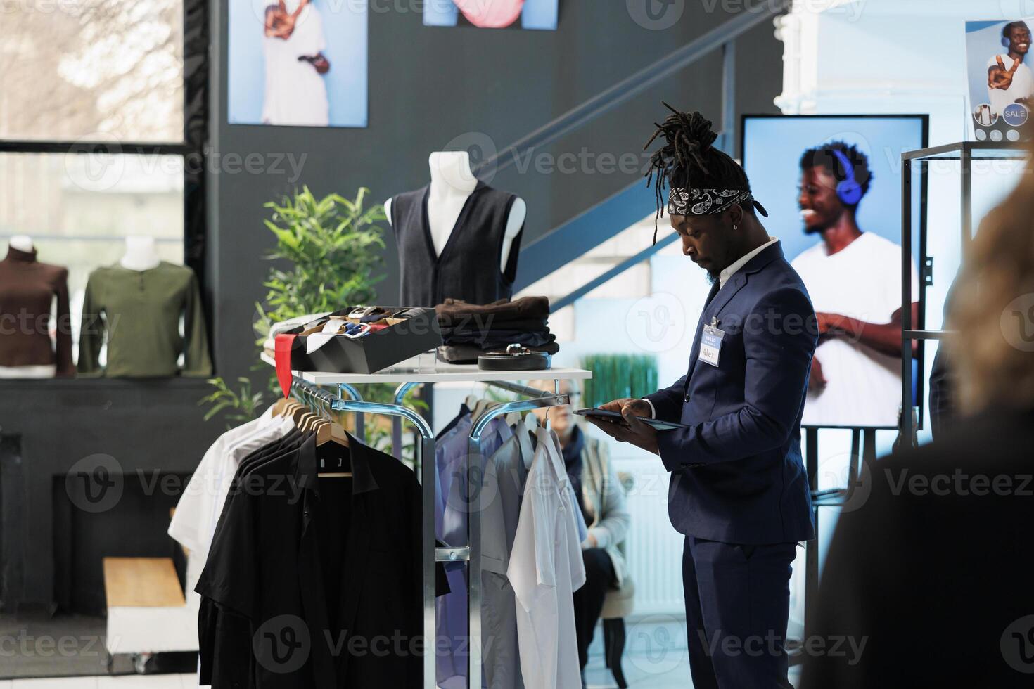 African american employee checking clothes inventory on tablet computer, looking at fashionable merchandise on hangers. Showroom worker checking discount list on device in shopping centre photo