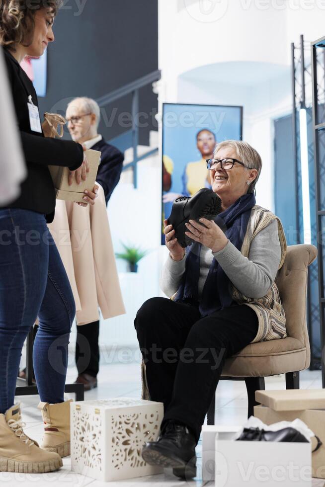 Senior woman trying fashionable black shoes, shopping for formal wear in clothing store. Elderly customer buying stylish clothes and merchandise in modern boutique. Commercial activity concept photo