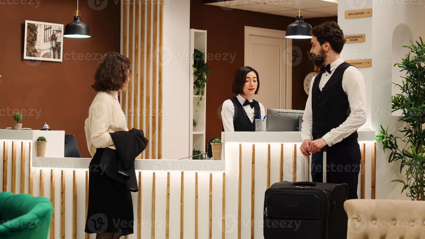 Guest arrives in hotel lobby, helped by bellboy with luggage and by asian receptionist with accommodation registration. Teamworking friendly helpful resort staff assisting tourist during check in photo