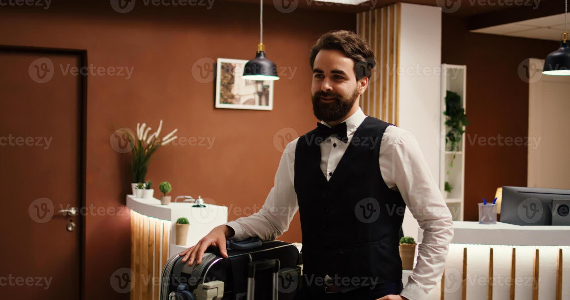 panorámica Disparo de contento sonriente profesional botones empleado en formal atuendo con equipaje carretilla en frente de cheque en recepción. alegre hotel trabajador que lleva equipaje en recurso vestíbulo foto