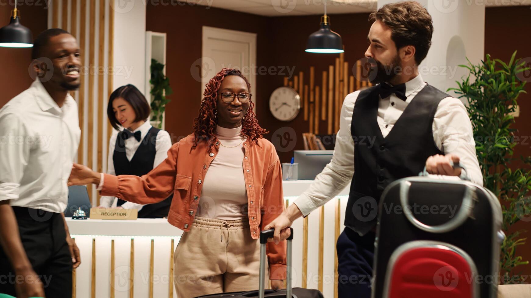 Happy african american tourists tipping helpful bellboy carrying their luggage in resort lobby. Cheerful guests checking out from hotel after handing over room access key card to friendly receptionist photo