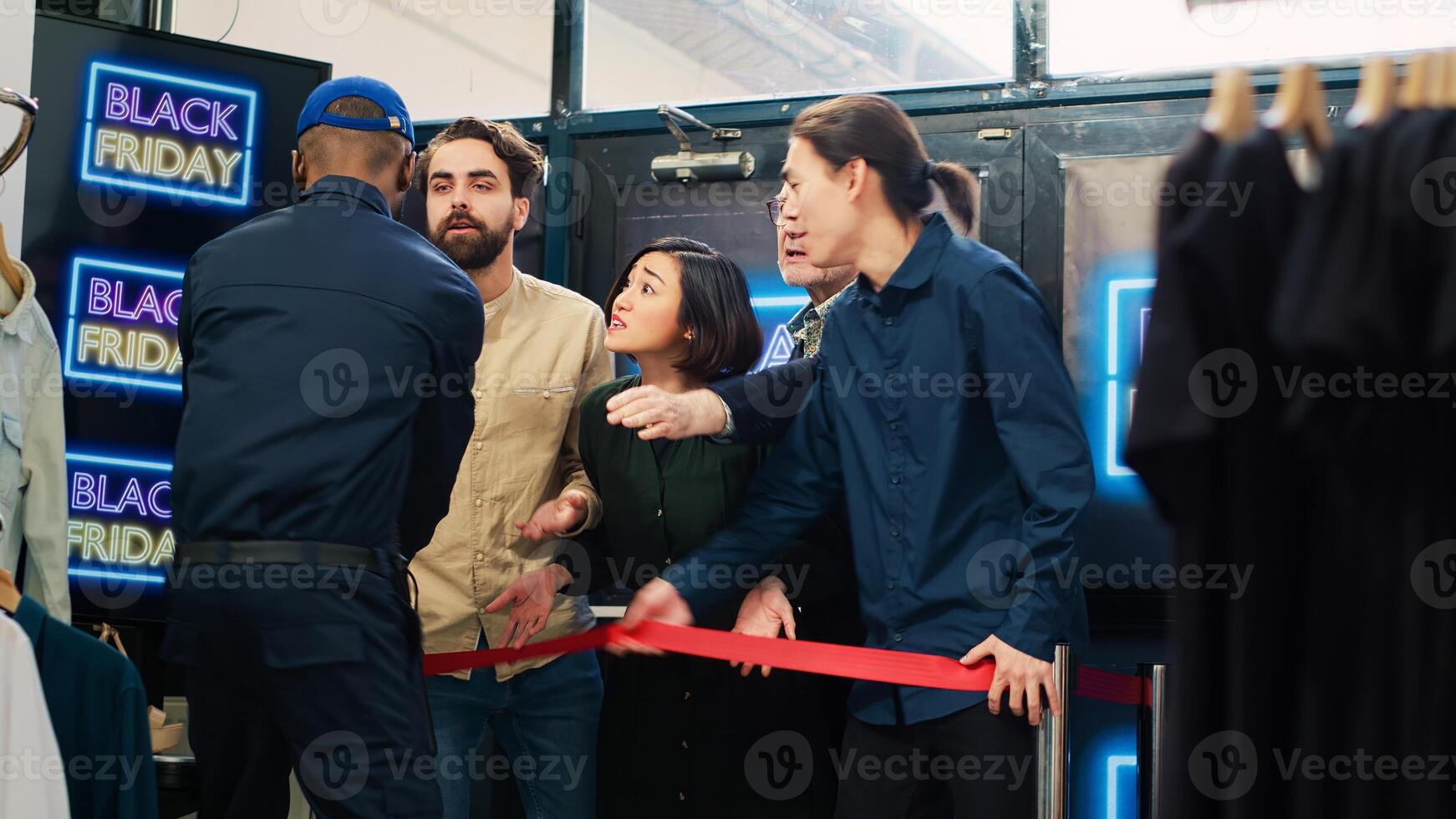 Customers pushing red tape and entering clothing store, looking to buy clothes on black friday sale. Security guard working under pressure with crowd of obsessed aggressive shoppers. Handheld shot. photo