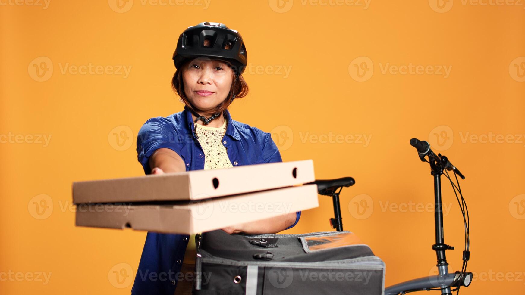 Courier woman isolated over orange studio background bringing pizza to customer. BIPOC bike rider delivering order to client, taking fast food boxes out of thermal backpack photo