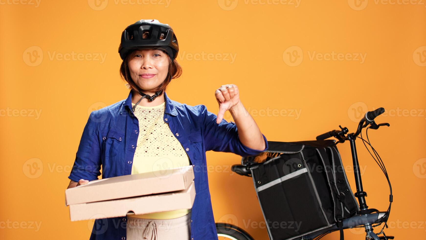 Portrait of professional courier holding fast food pizza boxes, doing dissaproving action. Bike rider showing thumbs down sign while delivering takeaway meal, isolated over studio background photo