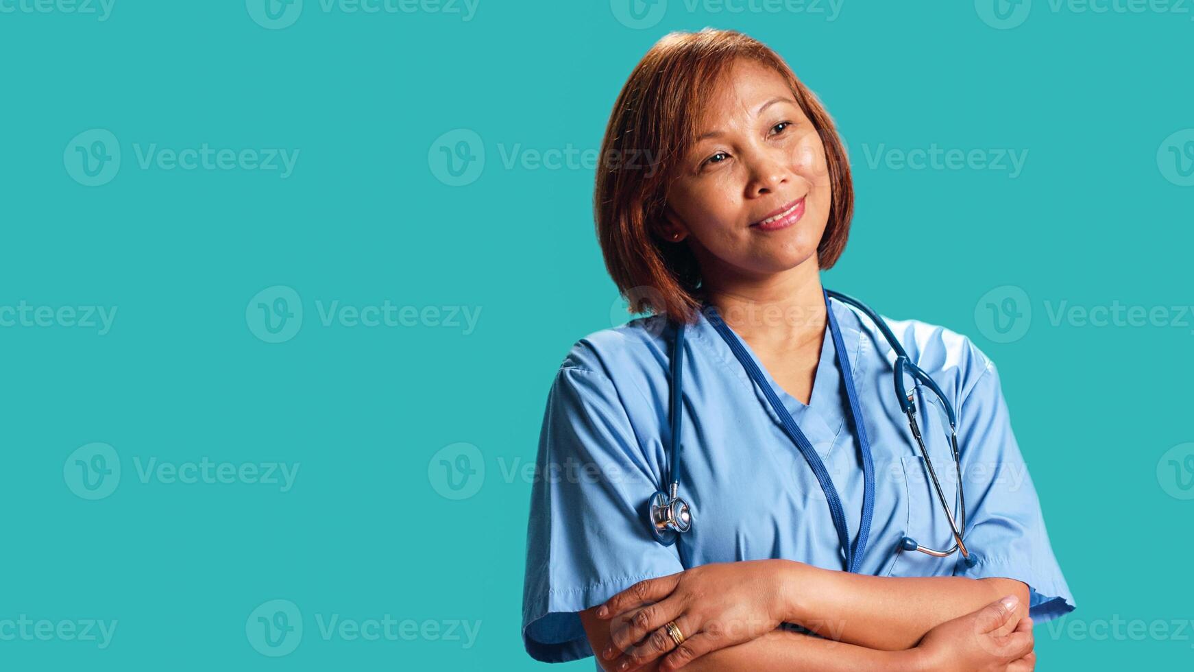 Joyful experienced asian nurse folding arms, smiling while looking at camera. Portrait of happy healthcare expert wearing protective scrubs, isolated over blue studio background photo