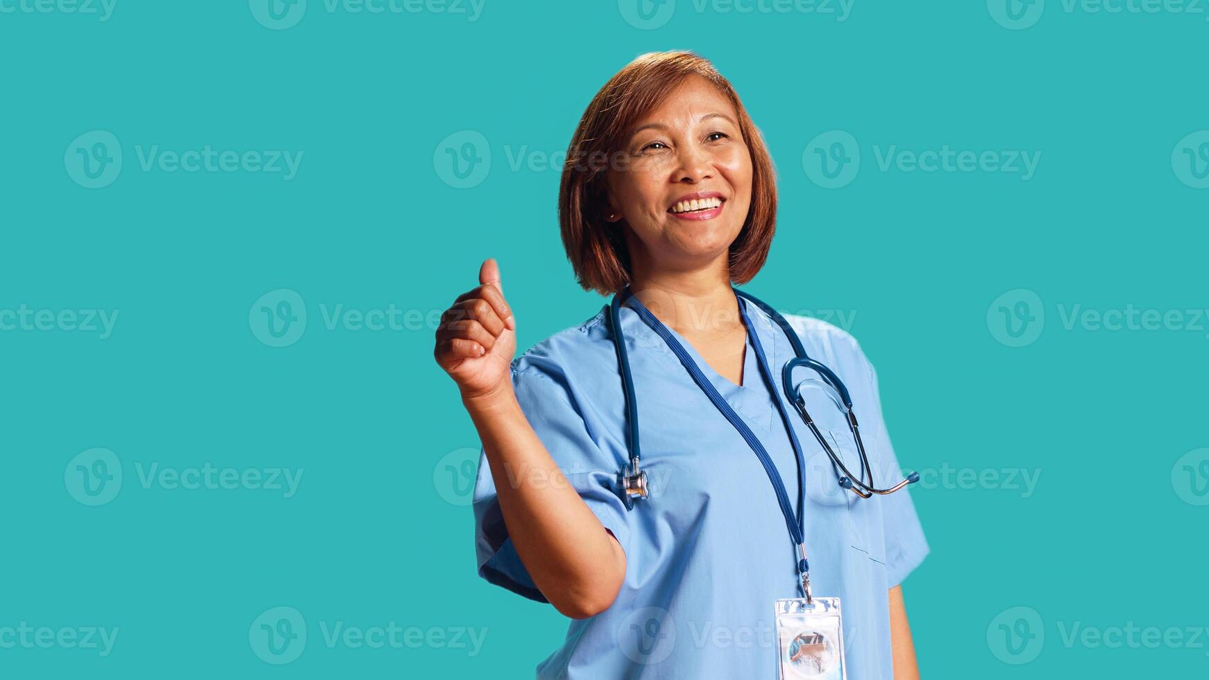 Upbeat expert BIPOC nurse showing thumbs up signs while at work, isolated over studio background. Close up shot of happy asian healthcare specialist doing approval gestures photo