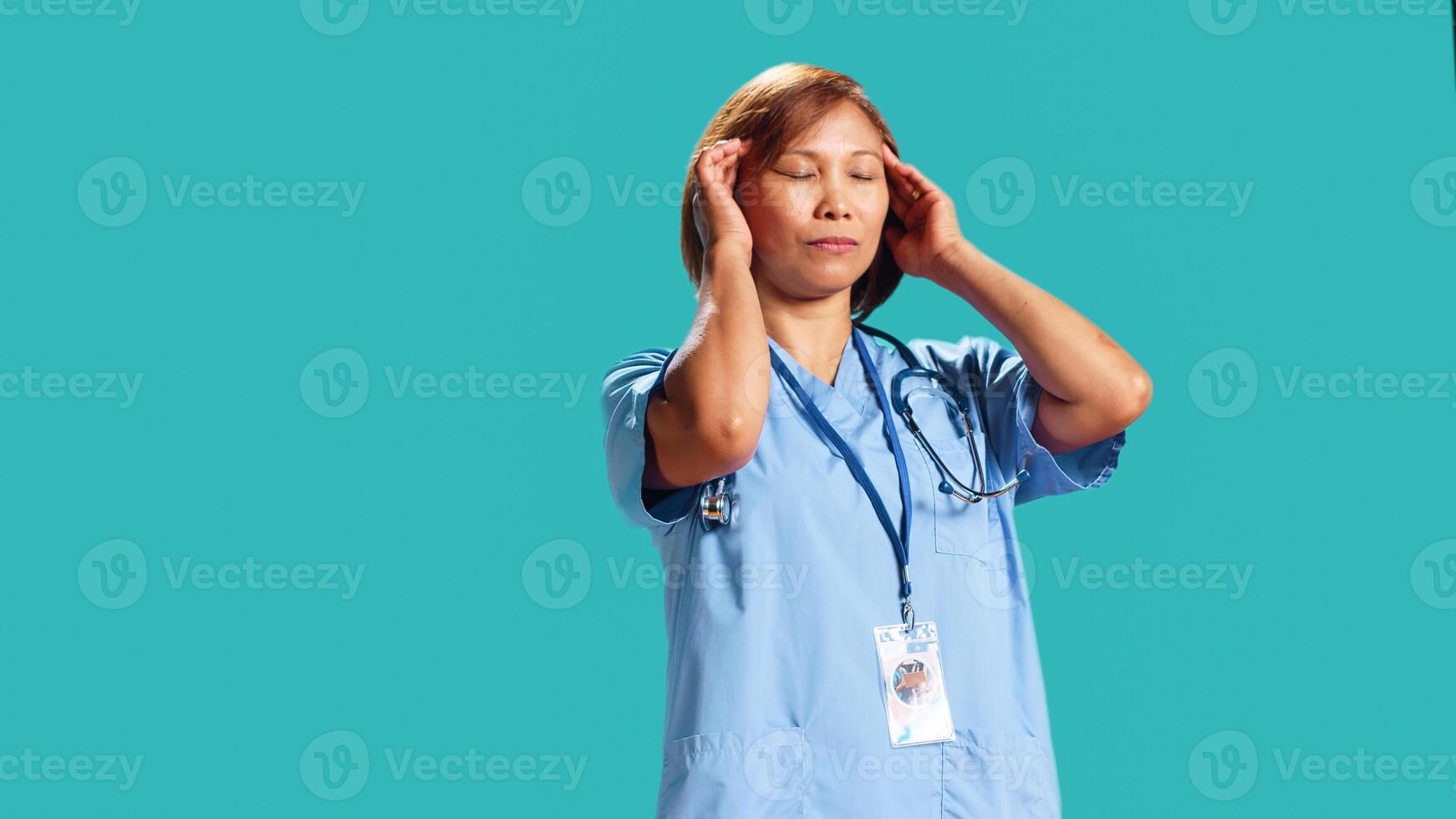 Fatigued nurse suffering from migraine pain after tiresome day at work. Hospital worker isolated over studio background affected by distressing headache during demanding job shift, close up photo