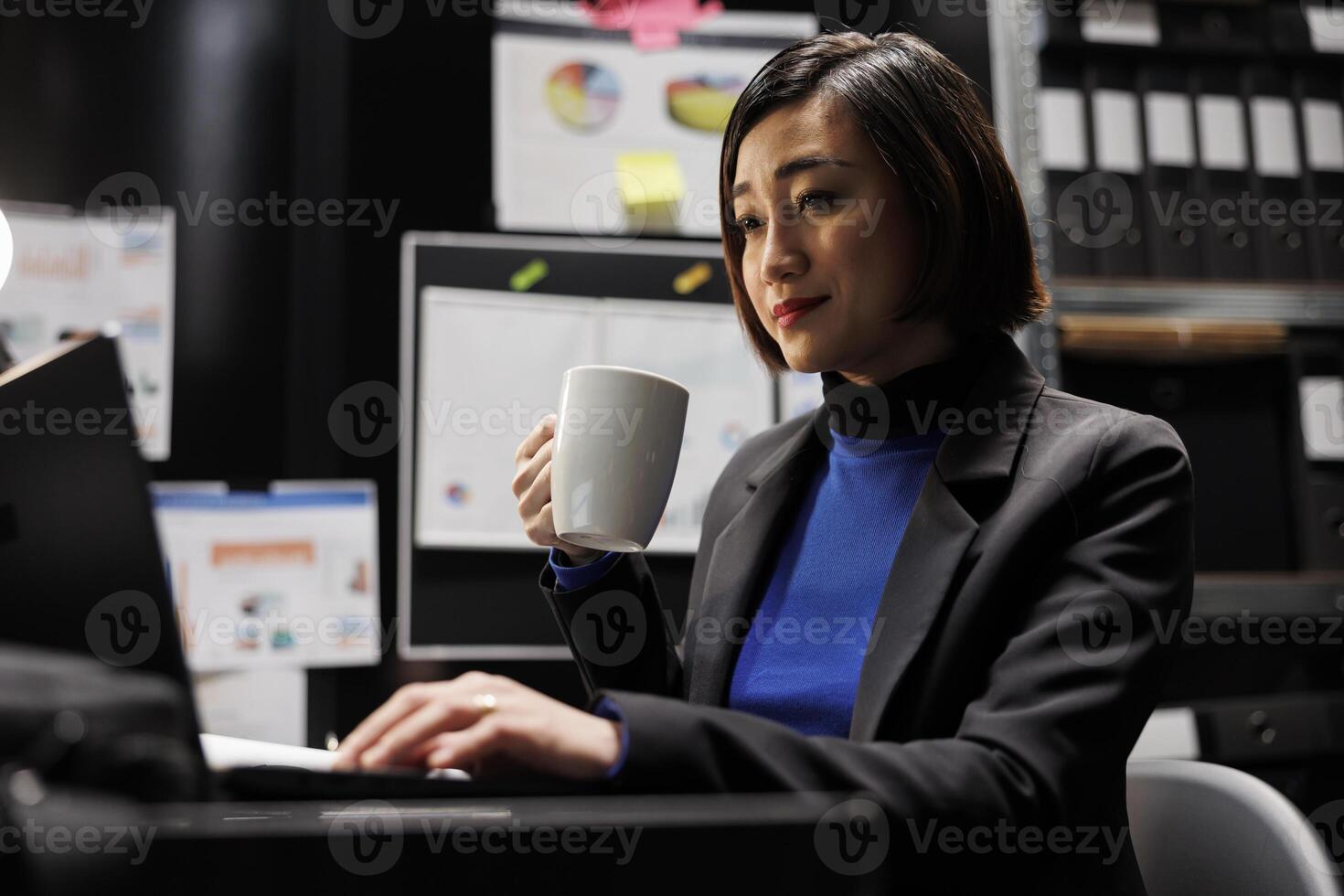 Cheerful bookkeeping businesswoman manager enjoying cup of coffee and imputing budget plan data on laptop in business office. Asian employee in file cabinet room with folders on bookshelves photo