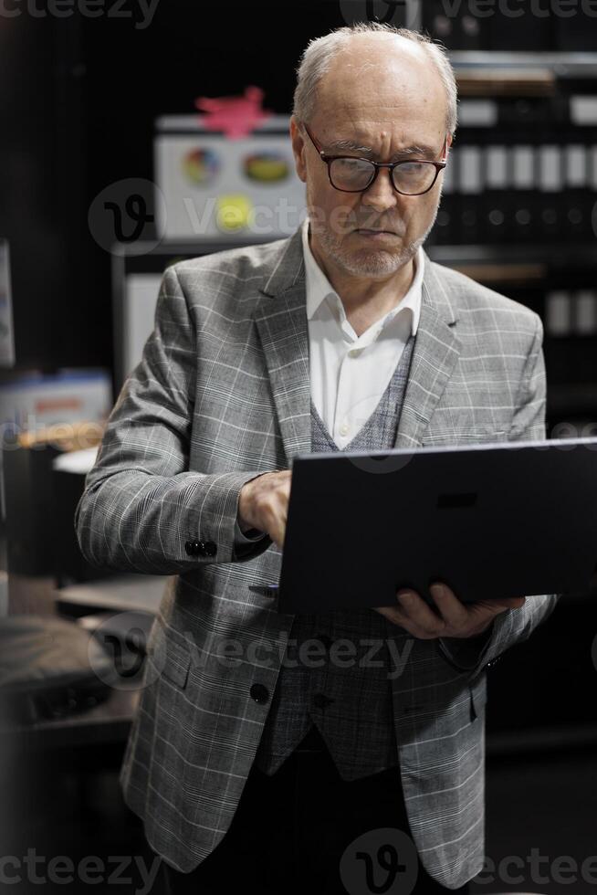 Old bookkeeping businessman in file cabinet office checking accounting analytical data on laptop. Administration elderly executive in business company repository filled with paperwork folders photo