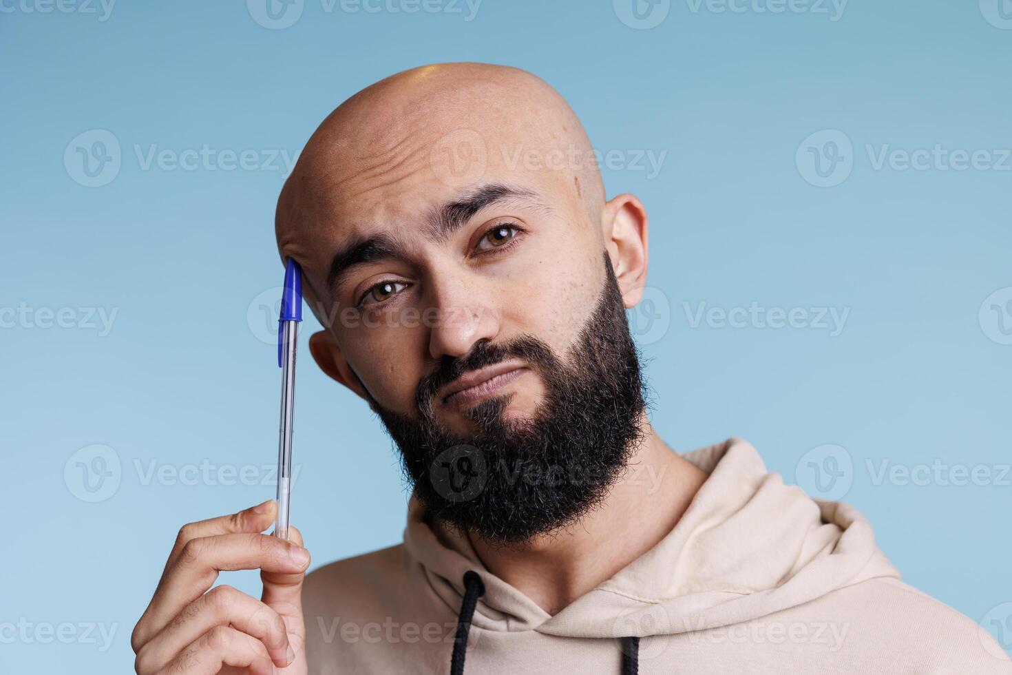 Puzzled arab man rubbing head with pen while thinking portrait. Young bald person in thought planning and solving problem while looking at camera with unsure facial expression photo