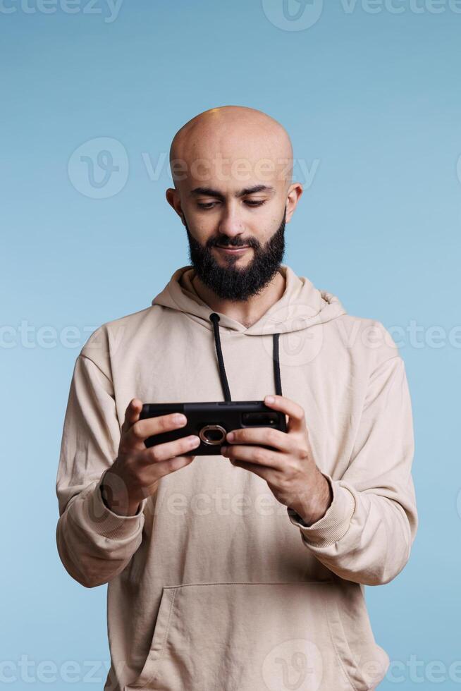 Arab man using smartphone application and playing video game during leisure activity. Young smiling person holding mobile phone in vertical mode and watching video online photo