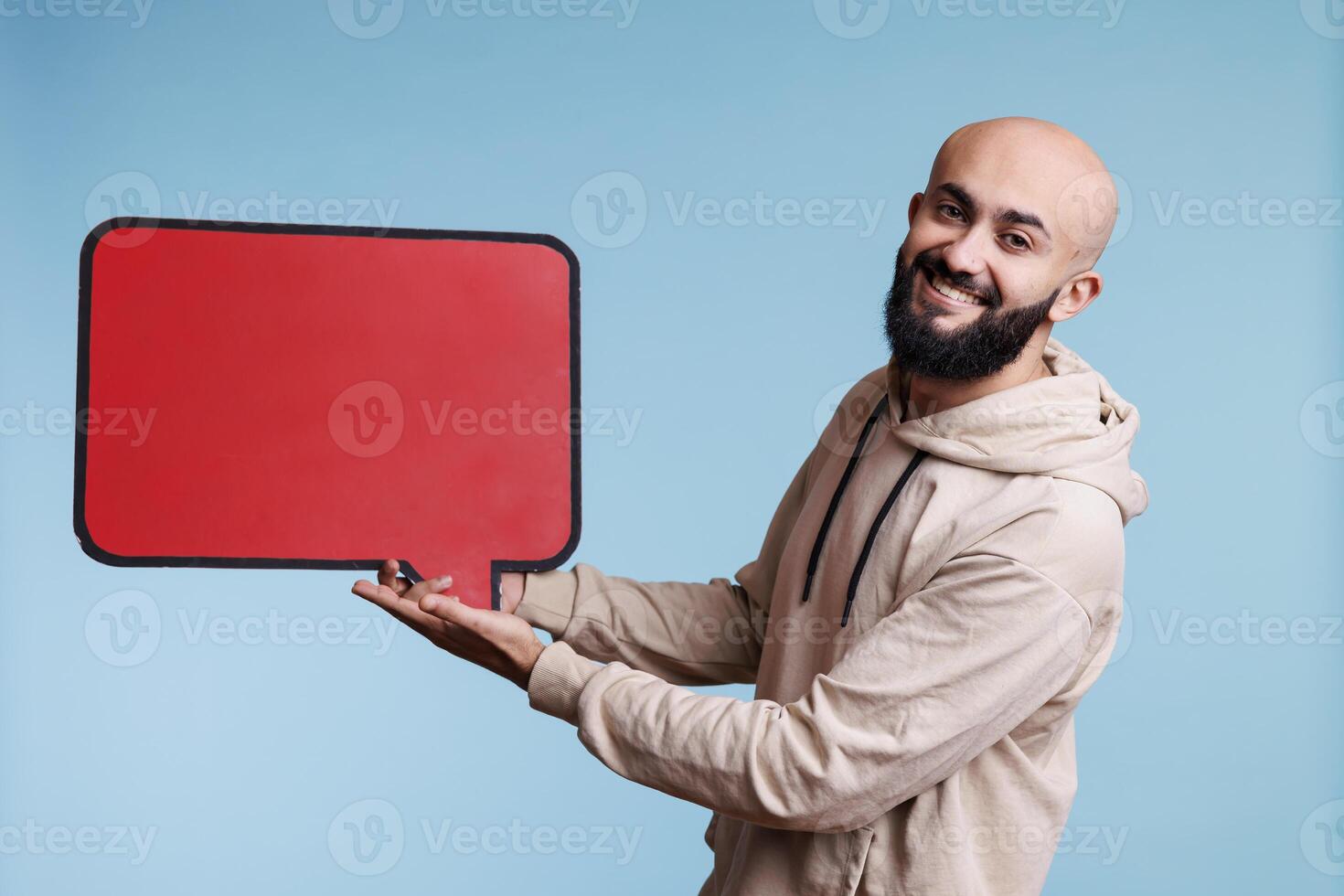 Arab man holding blank red speech frame product marketing mock up and looking at camera. Smiling carefree person showing empty message frame for advertising studio portrait photo