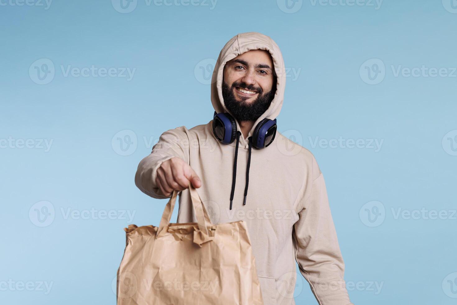 Cheerful smiling arab man in hood giving paper bag with take out food portrait. Restaurant delivery service courier holding package with takeaway lunch and looking at camera photo