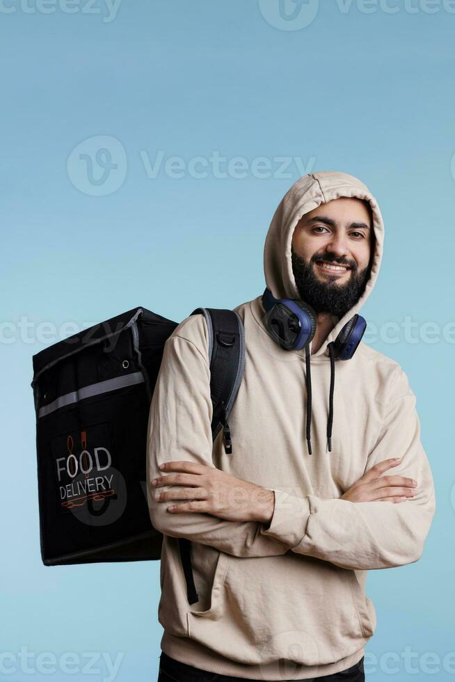 sonriente árabe hombre posando con comida entrega Servicio mochila y mirando a cámara con alegre expresión. contento repartidor en pie con térmico bolso con restaurante almuerzo retrato foto