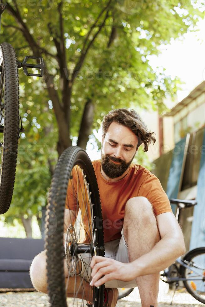 Active and committed man crouching to repair bike wheel rim in home yard using professional tool. Healthy male cyclist working on dismantled bicycle tire outside using specialized wrench. photo