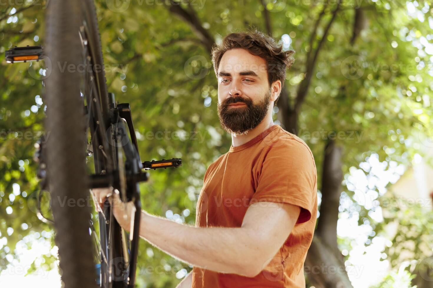 Young sports enthusiast man checks bicycle parts in yard for damage that can be fixed with tools. Active male cyclist repairing damaged wheel and chain outside for summertime pleasure cycling. photo